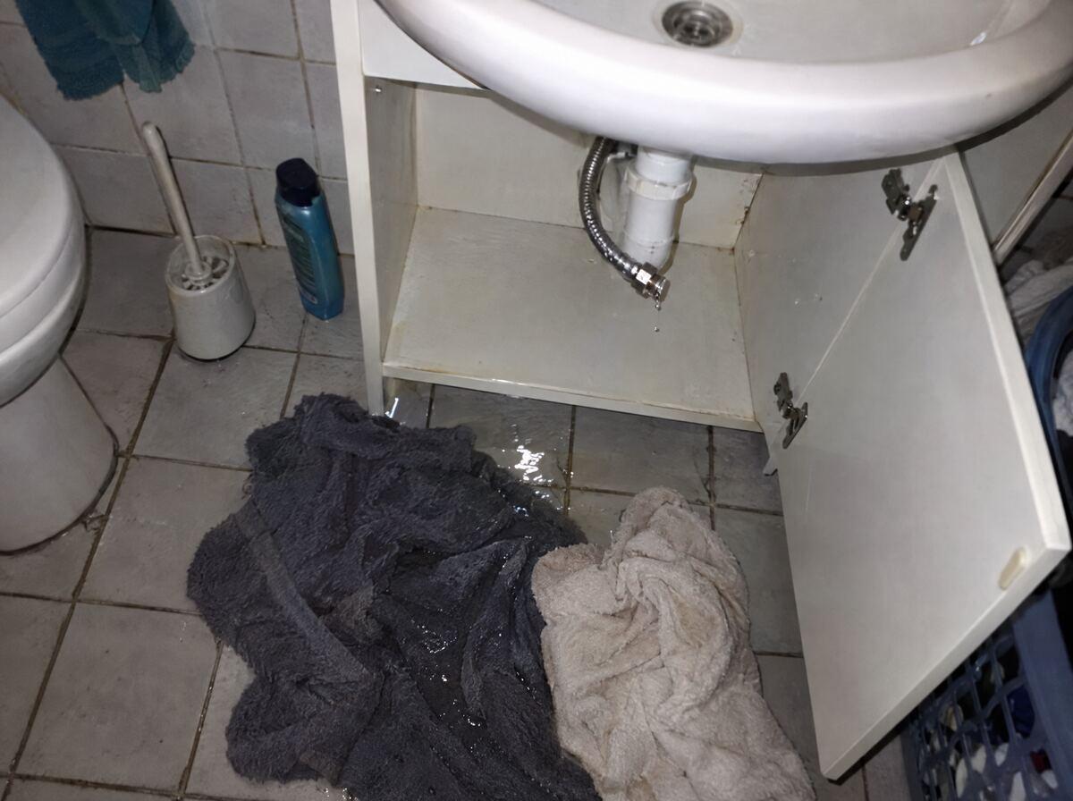 Water op de badkamervloer onder een wastafel, natte handdoeken erop
